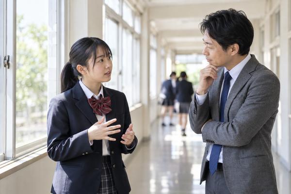 休み時間の廊下で、授業中に困っている場面を先生へ具体的に伝えている制服の生徒