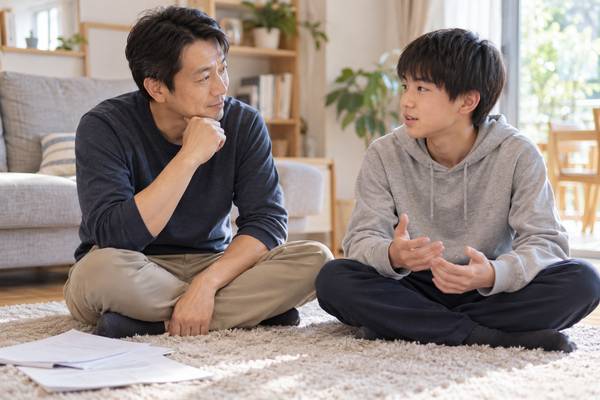 明るいリビングで、父親が中学生の息子の話に耳を傾け、返却された答案は脇に置かれている様子