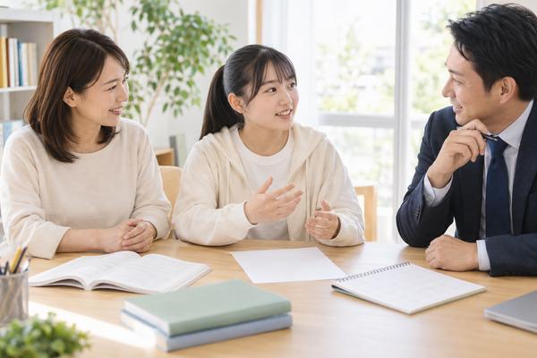 明るい相談室で、高校生が答案と教材と1週間の記録を広げて自分から話し、保護者と先生が耳を傾けている様子