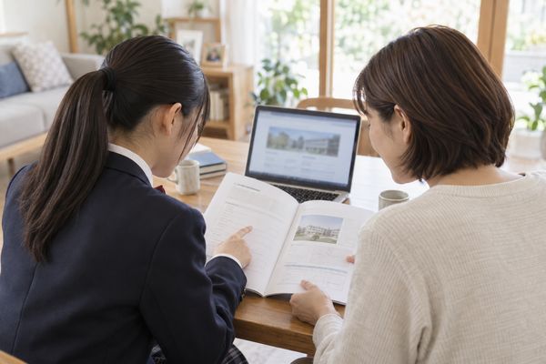 明るいリビングで、大学の募集要項とノートパソコンの公式サイトを保護者と一緒に確認する制服姿の高校生