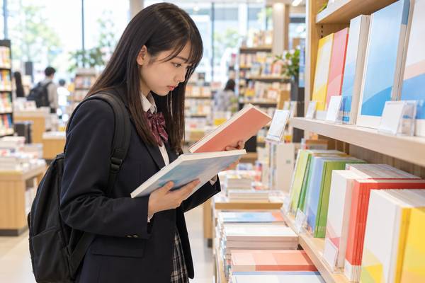 書店の棚で2冊の参考書を見比べ、どちらの版かを確かめている制服の高校生
